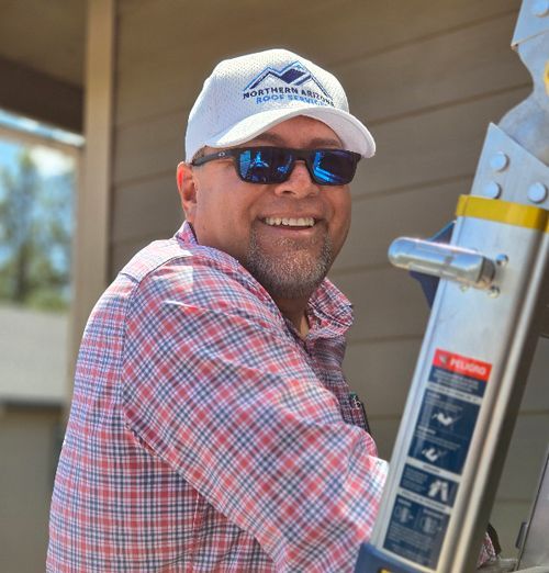 Man in plaid shirt and sunglasses smiles, holding onto a ladder outdoors, wearing a cap.
