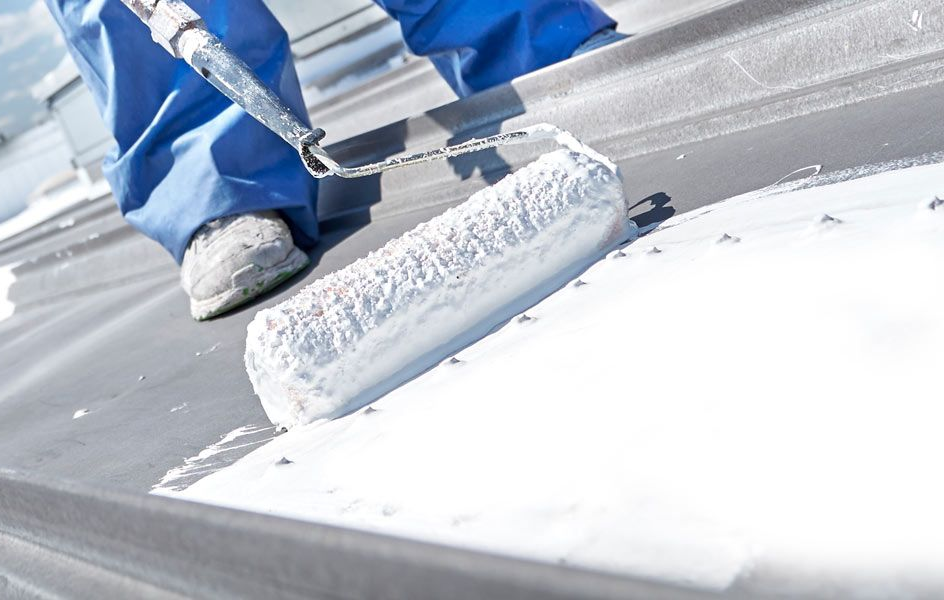 A worker applying sealant to a flat roof with a torch, wearing gloves, blue jeans, and an orange plaid shirt.