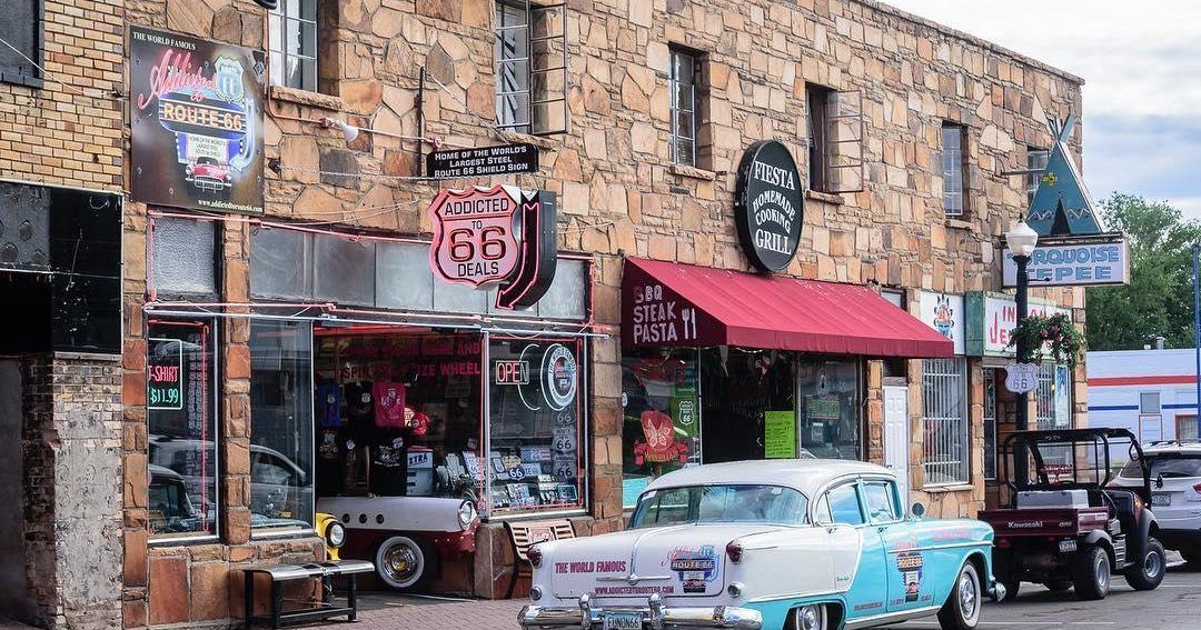Shops with retro Route 66 signage; classic cars parked in front of a stone-clad building.