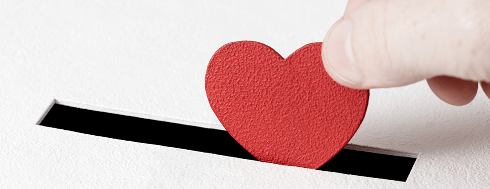 Hand inserting a red heart into a black slot on a white surface, symbolizing giving or donation.