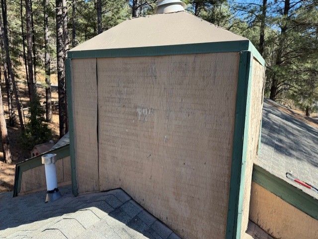 Chimney on a roof with green trim and beige panels, surrounded by trees.