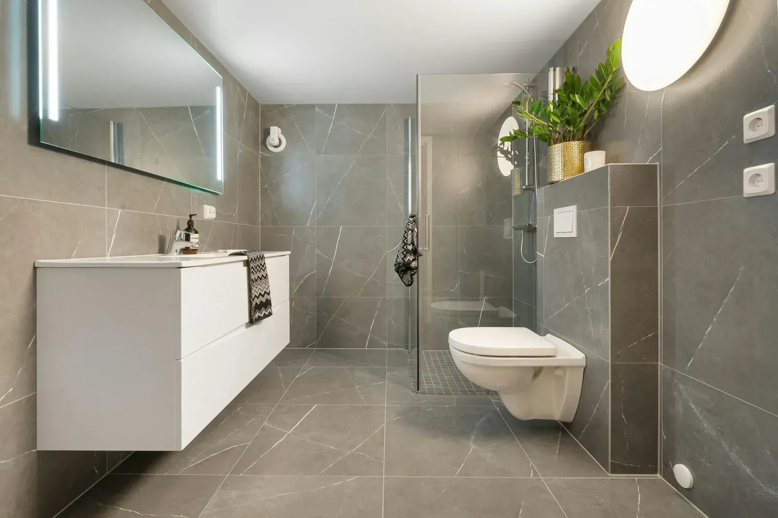 Modern gray bathroom with floating white vanity, toilet, glass shower, and potted plants.