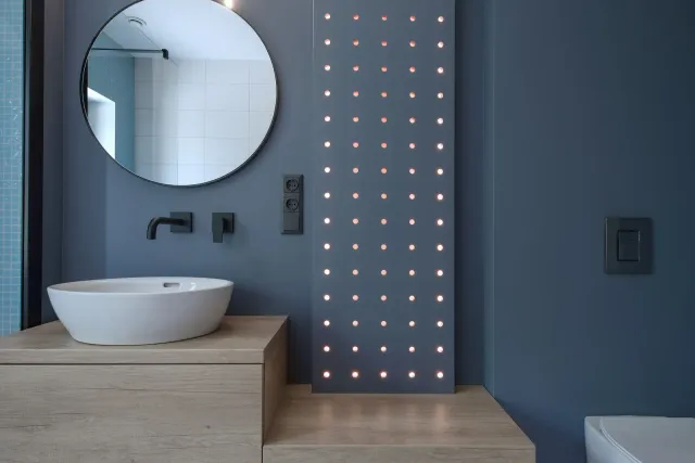 Modern bathroom with a blue wall, round mirror, and a white sink on a wooden counter.
