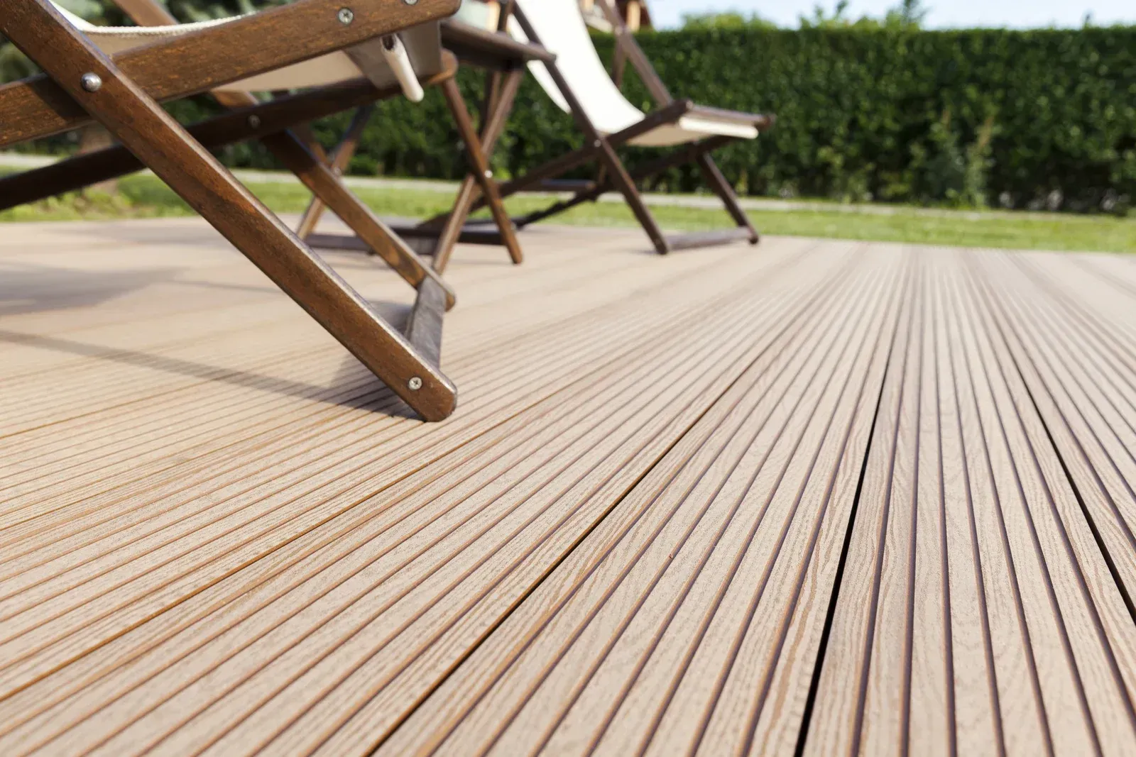 Two wooden chairs on a brown, grooved wooden deck, with a green hedge and lawn in the background.