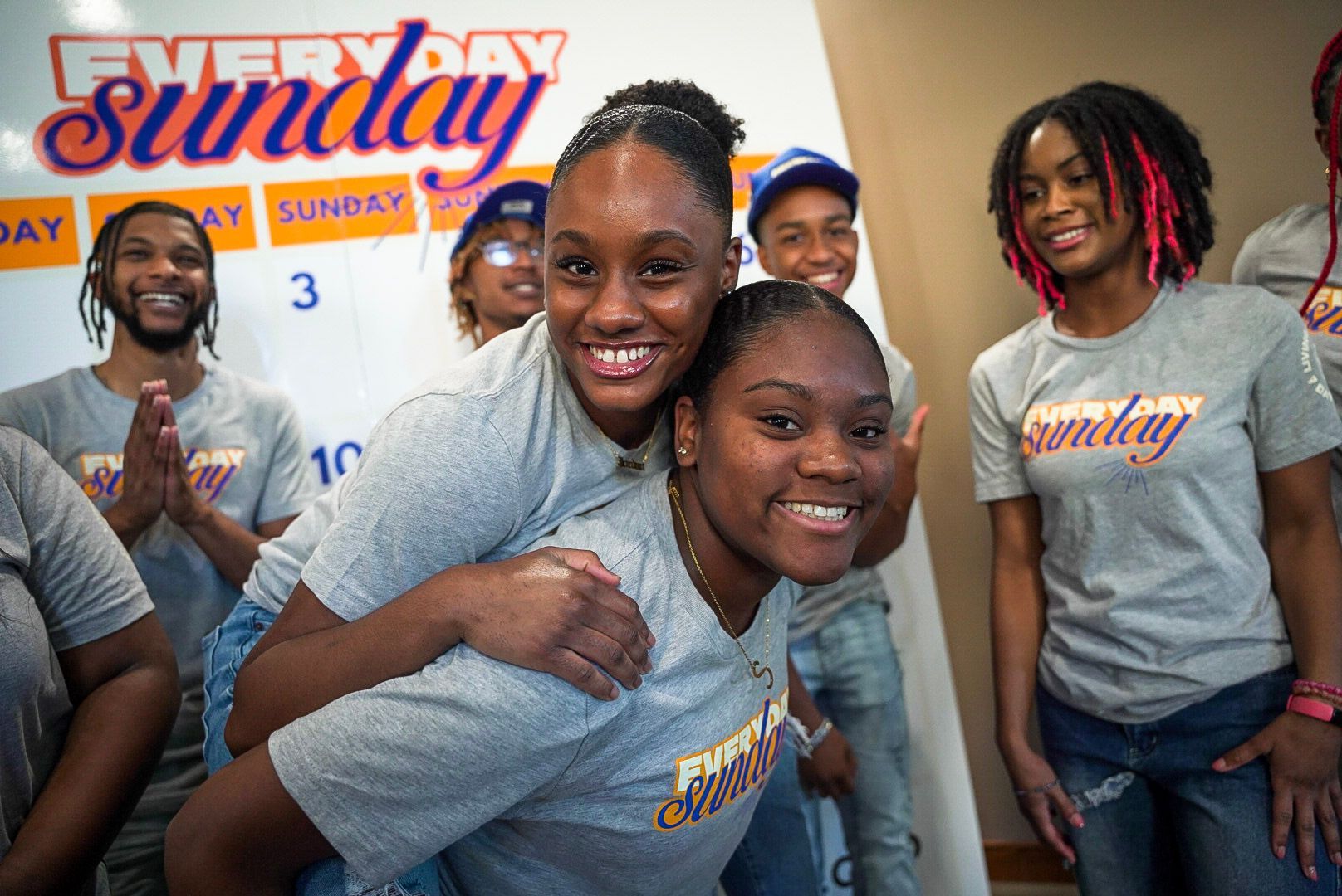 Two smiling women in grey shirts; one gives the other a piggyback ride. Group of smiling people in background.