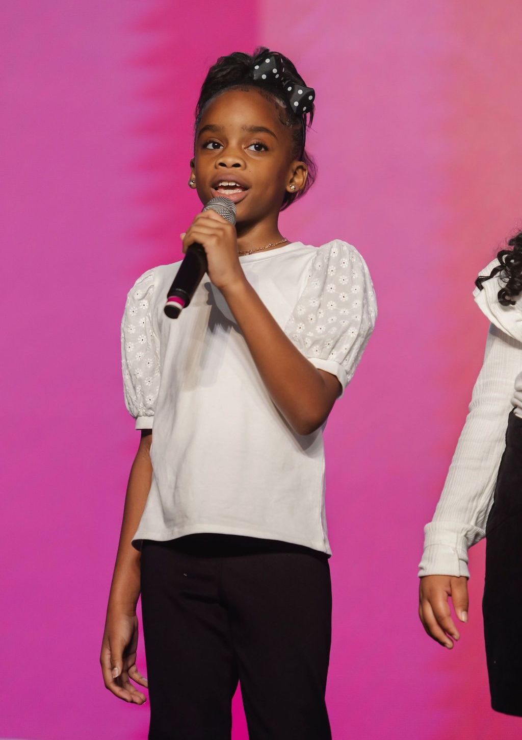 Young Black girl singing into a microphone on a pink stage, wearing a white eyelet top and black pants.