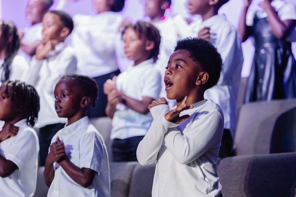 Children's choir singing with hands on their chests in a brightly lit performance hall.