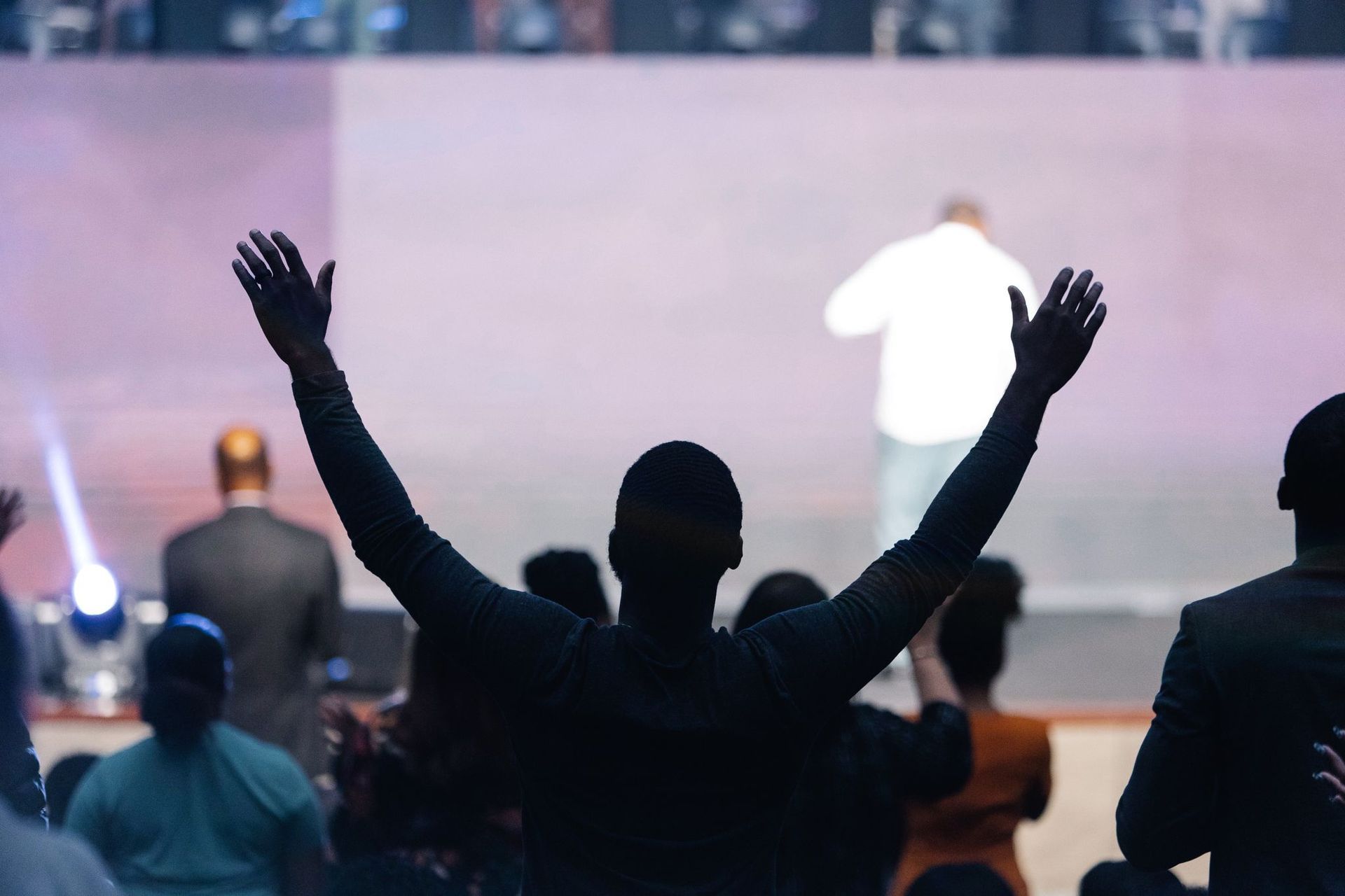 Silhouette of person with arms raised in worship, facing a stage with a person, during a service.