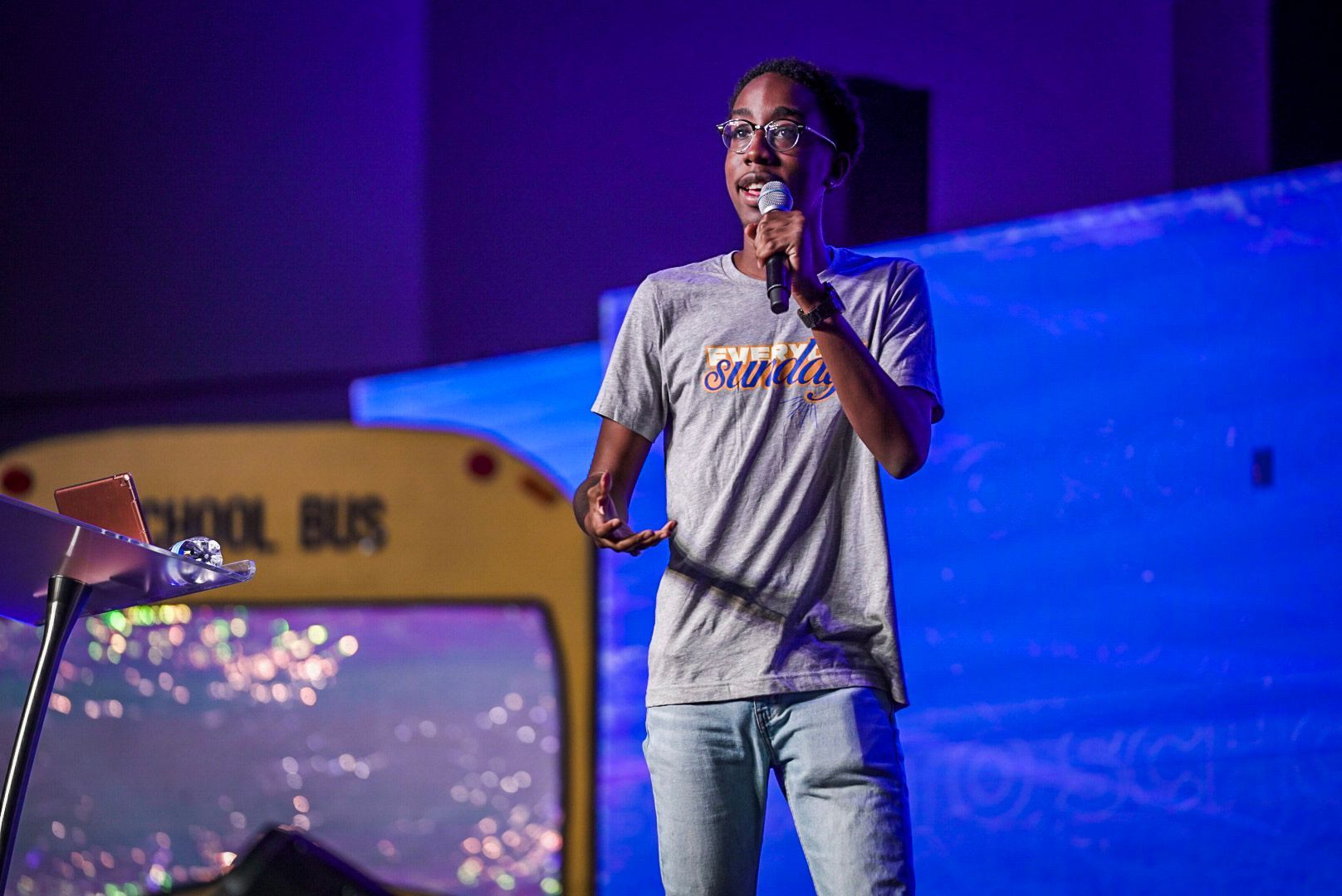Person speaking into a microphone on a stage with a school bus backdrop and blue lighting.