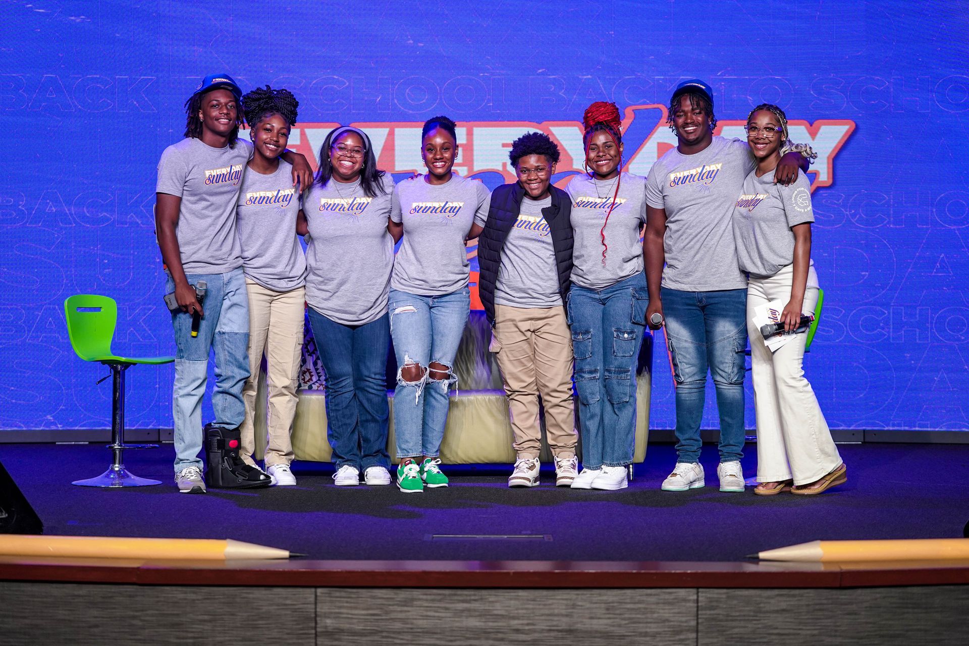 Group of diverse people in grey shirts on stage, smiling. Blue background, microphones present.