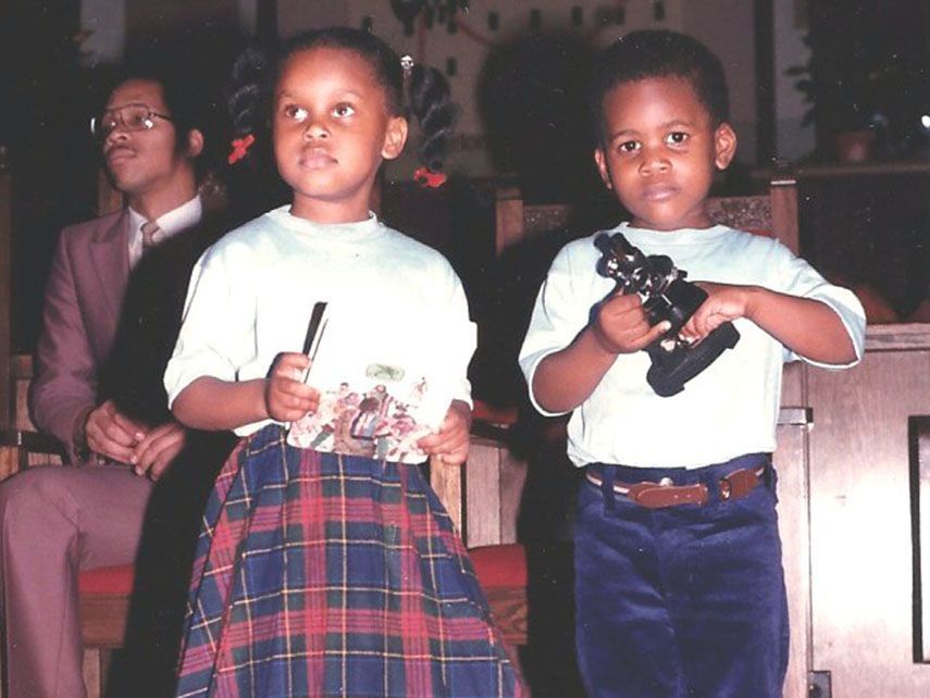Two Black children hold objects; a girl in plaid skirt, a boy in blue pants; man in background.