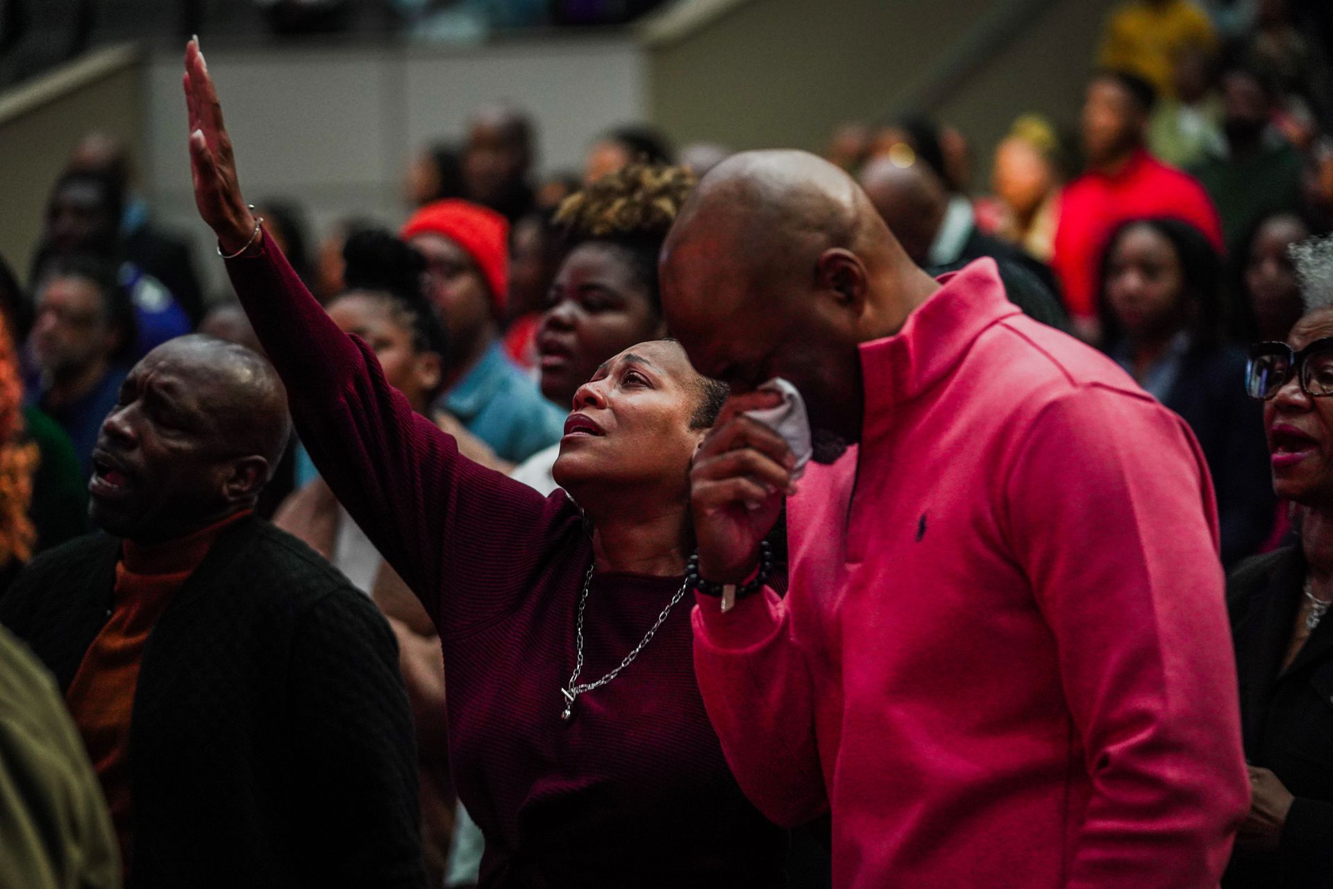 People in church, some with hands raised in worship, a woman in a maroon shirt has her hand raised high.