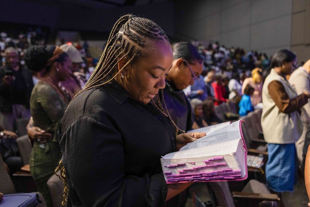 Woman with braids reads a book in a church, surrounded by people in rows of seats.