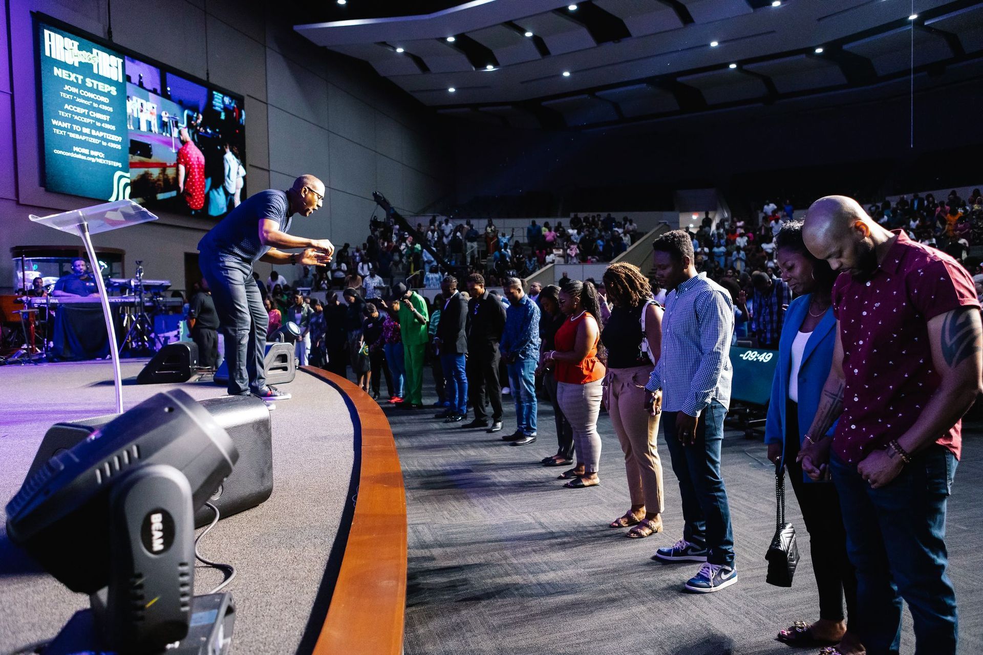 A man speaking on stage to a line of people in a large church. Audience in background.