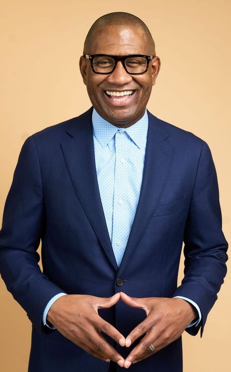Brian Carter navy blazer, light blue patterned shirt, hands clasped, against tan backdrop.
