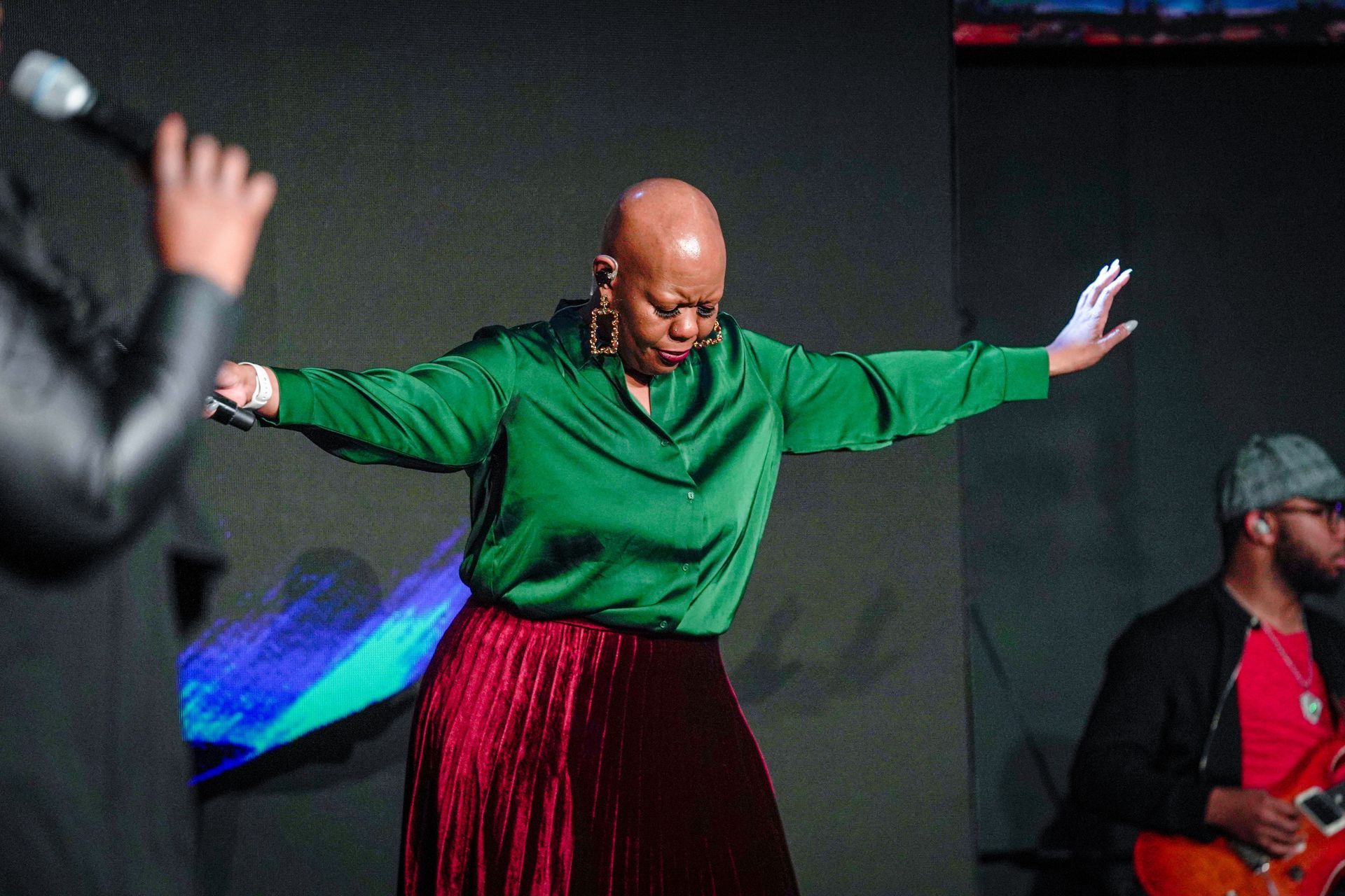 Woman with shaved head singing, arms outstretched, wearing a green shirt and red skirt on a stage.