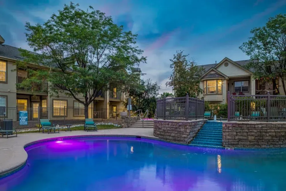 Apartment complex pool at dusk, lit with blue and purple lights. Buildings and trees surround the pool.