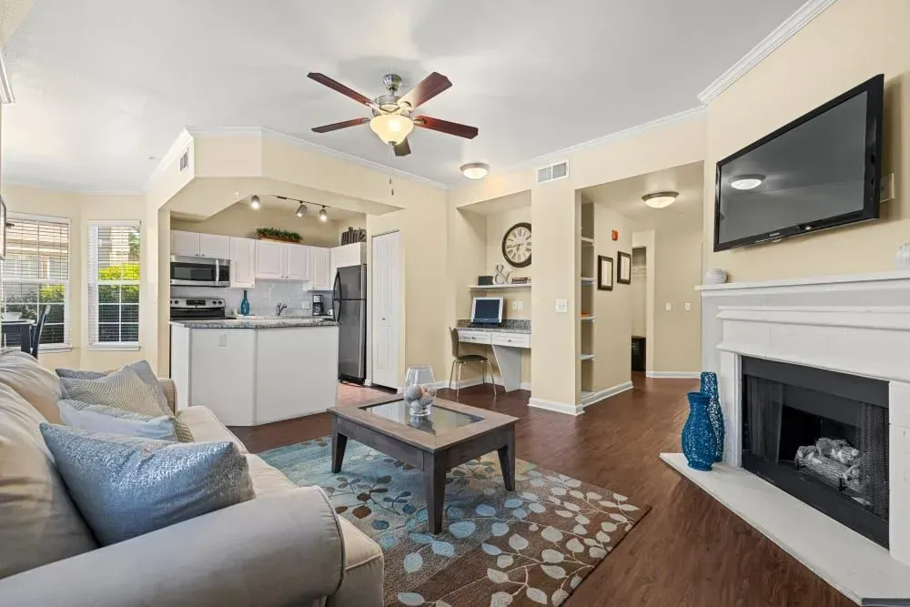 Cozy living room with open kitchen; cream-colored walls, a fireplace, and a mounted TV. A sofa faces the fireplace, and a desk is tucked in a nook.