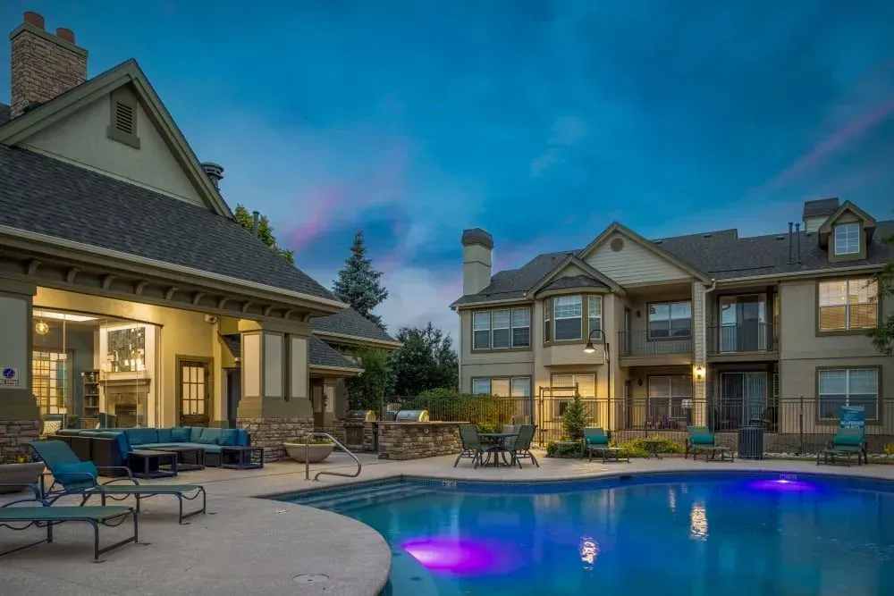 Nighttime exterior of a luxury apartment complex with a pool. Buildings have outdoor seating, a fireplace, and a gas grill around the illuminated pool.