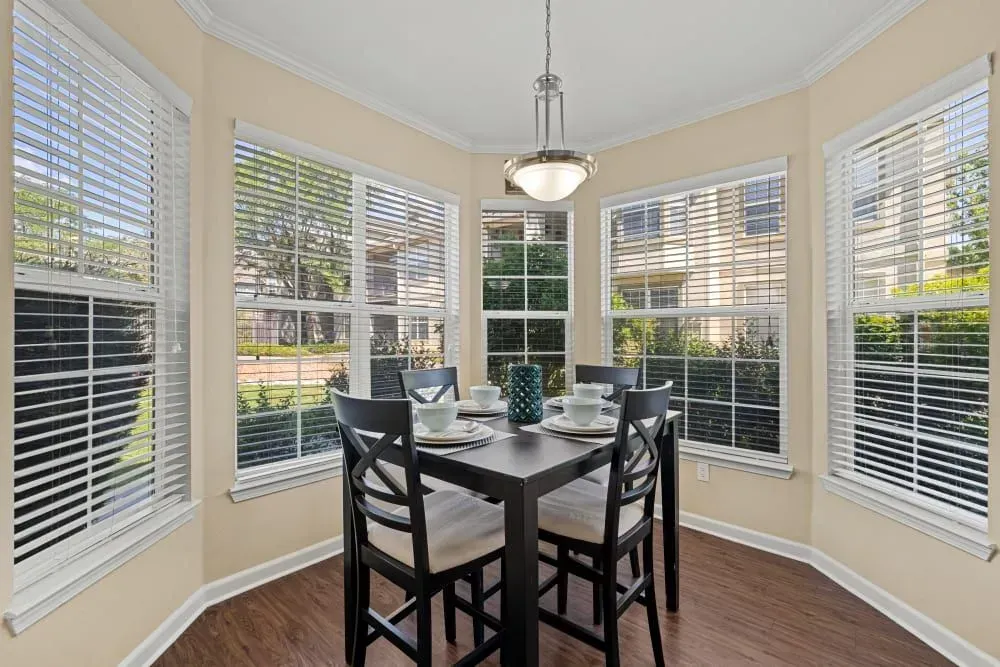 A dining area with a dark square table, chairs, and a hanging light. Large windows with blinds offer a view of the outside.