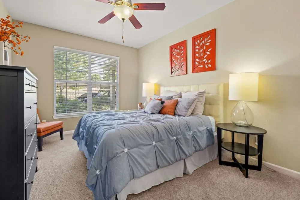 Bedroom with a bed, nightstand, dresser, and window. Blue and orange accents, beige walls and carpet.