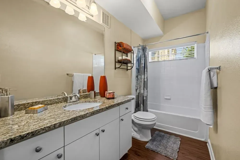 A bathroom with tan walls, a white tub/shower, and a white vanity with a granite countertop.