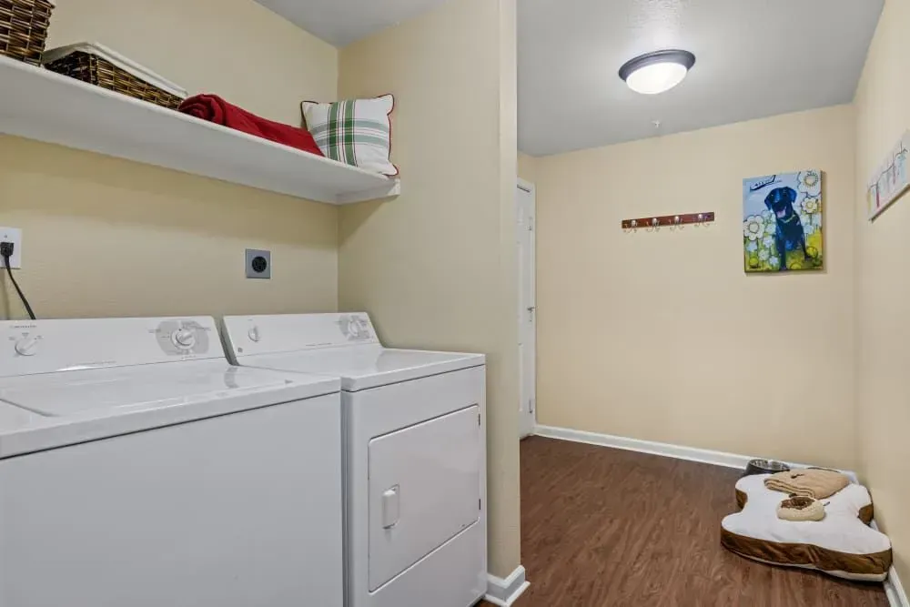 A laundry room with white washer and dryer, a shelf with linens, and a hallway leading to a door. Brown floor and beige walls.