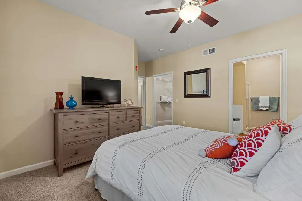 Bedroom with a light-colored bed, wooden dresser with TV, and a doorway to a bathroom.
