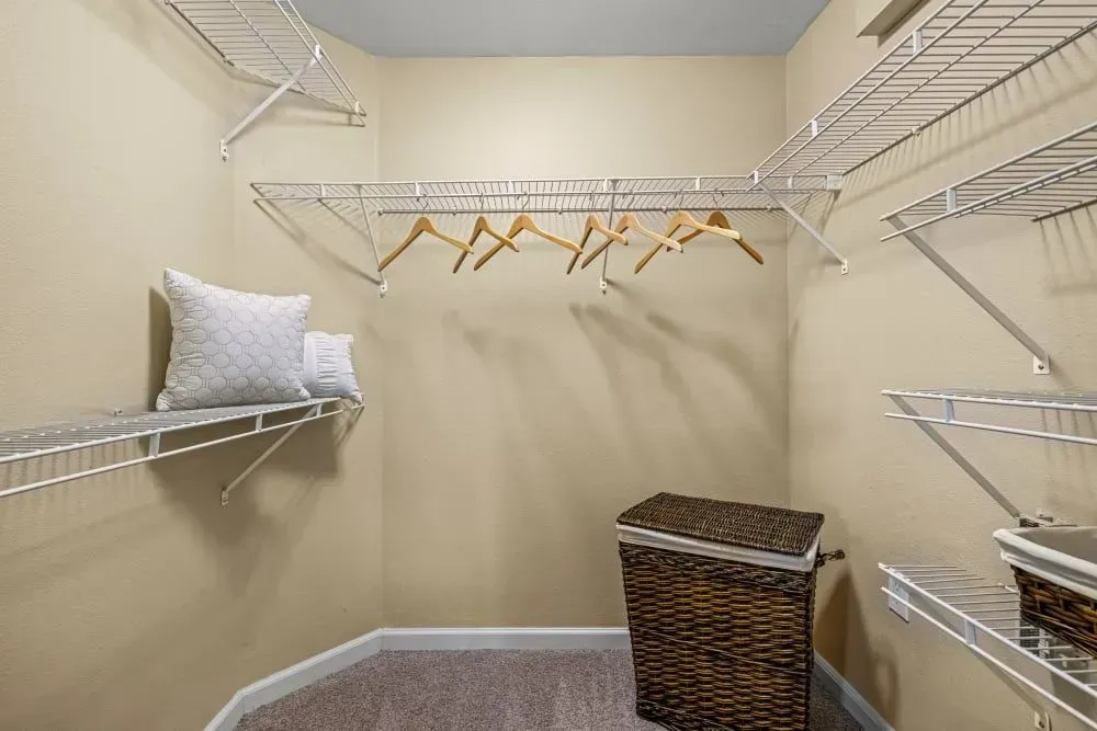Empty walk-in closet with wire shelves, beige walls, and a woven hamper. Wooden hangers hang from a rod.