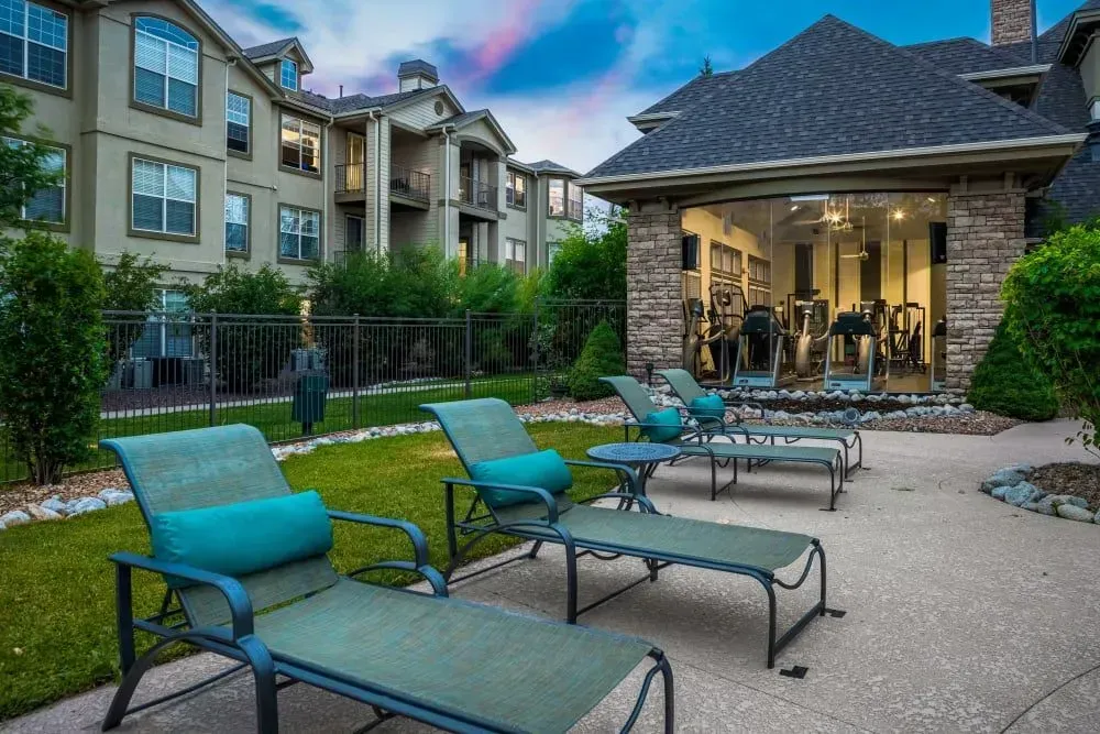 Lounge chairs on a patio overlook a gym with exercise equipment, set beside an apartment building under a twilight sky.