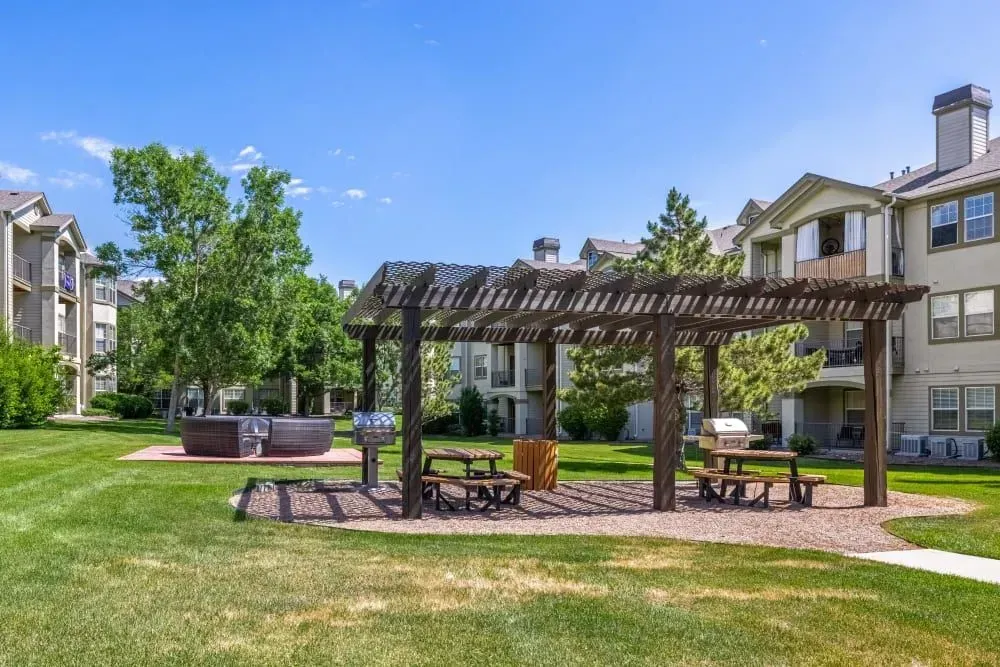 A grassy outdoor area with a pergola, picnic tables, and a grill, likely a residential amenity.
