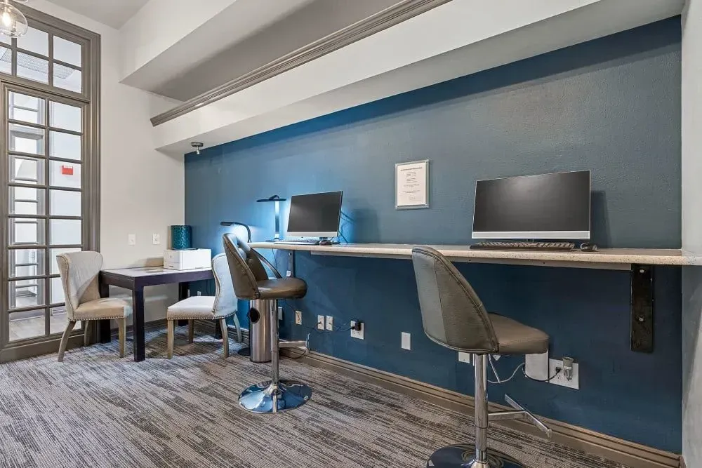A modern business center with a navy blue wall, computers, high-top chairs, and a small table near a large window.