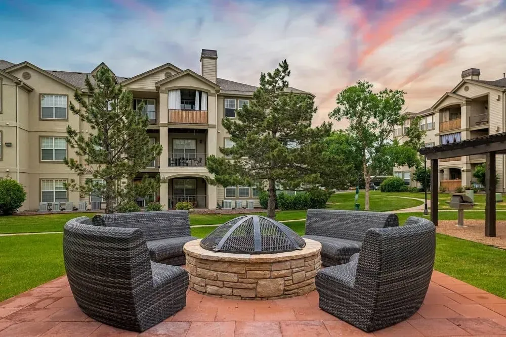 A patio with a stone fire pit and wicker chairs surrounded by an apartment complex, with a colorful sunset in the background.