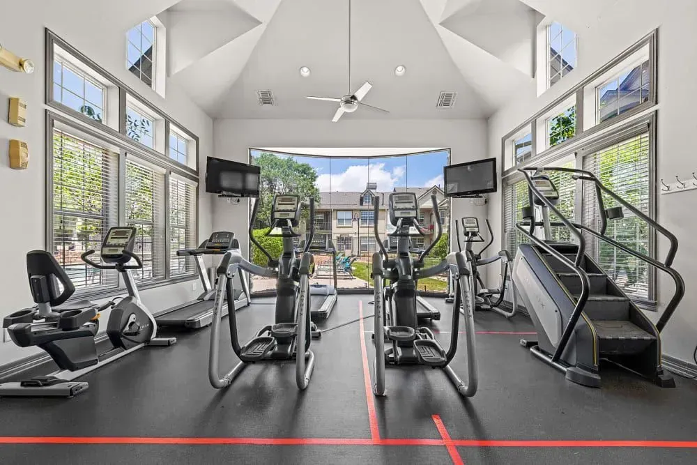Gym interior with exercise equipment, large windows, and two TVs. Includes treadmills, ellipticals, a stair climber, and exercise bikes.