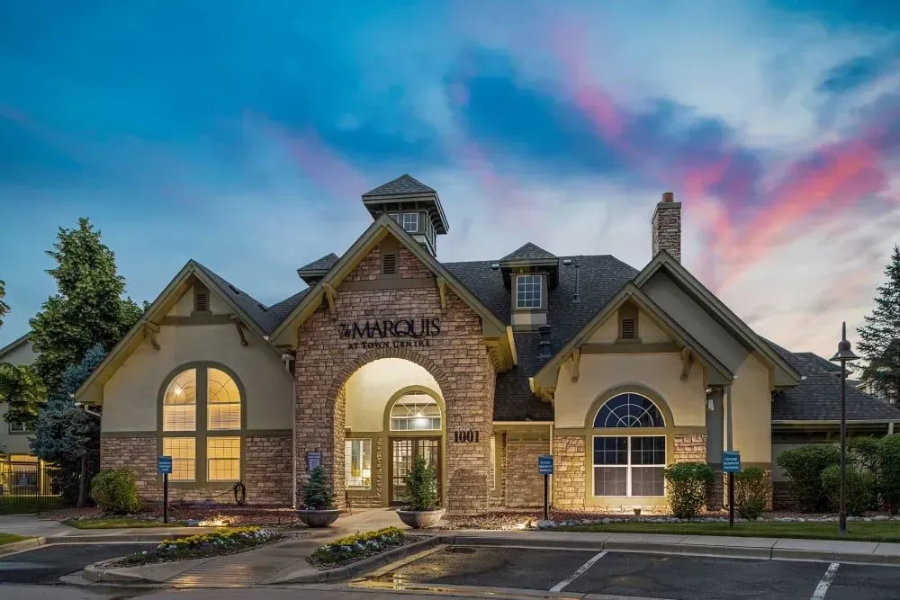 Marquis apartment building exterior with brick facade, arched entryway, and parking spaces in front. The sky has colorful sunset hues.