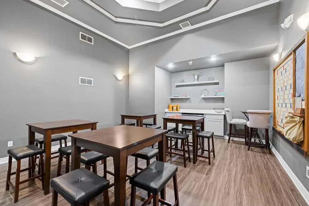 A community room with dark tables, black stools, a small kitchenette, and a decorative ceiling. Gray walls and wood-look floors.