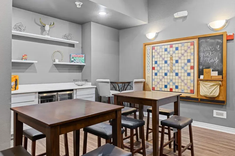 A game room with tables, stools, a large Scrabble board, and shelves with decorative items against gray walls.