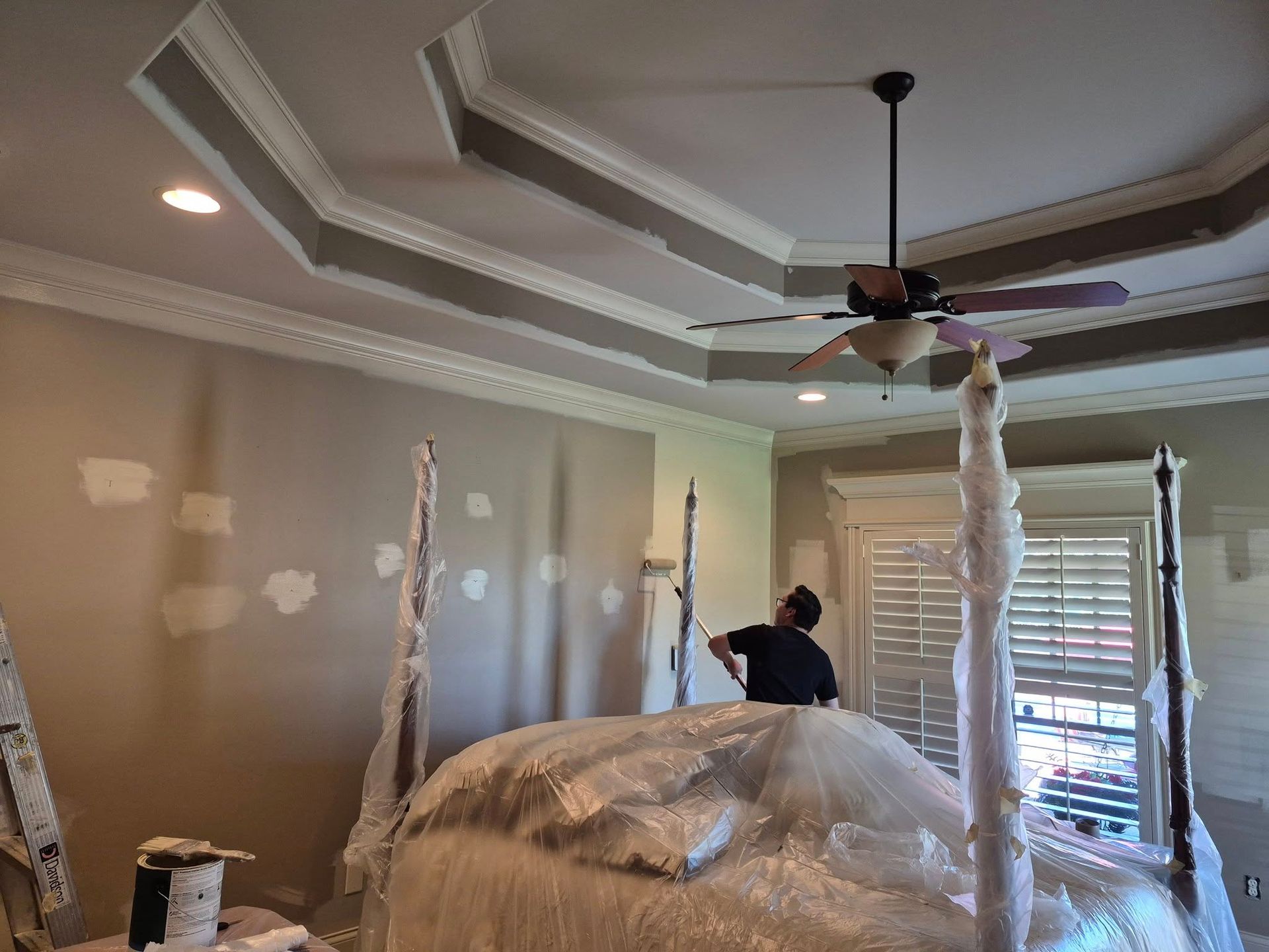 A man is painting a four poster bed in a bedroom with a ceiling fan.