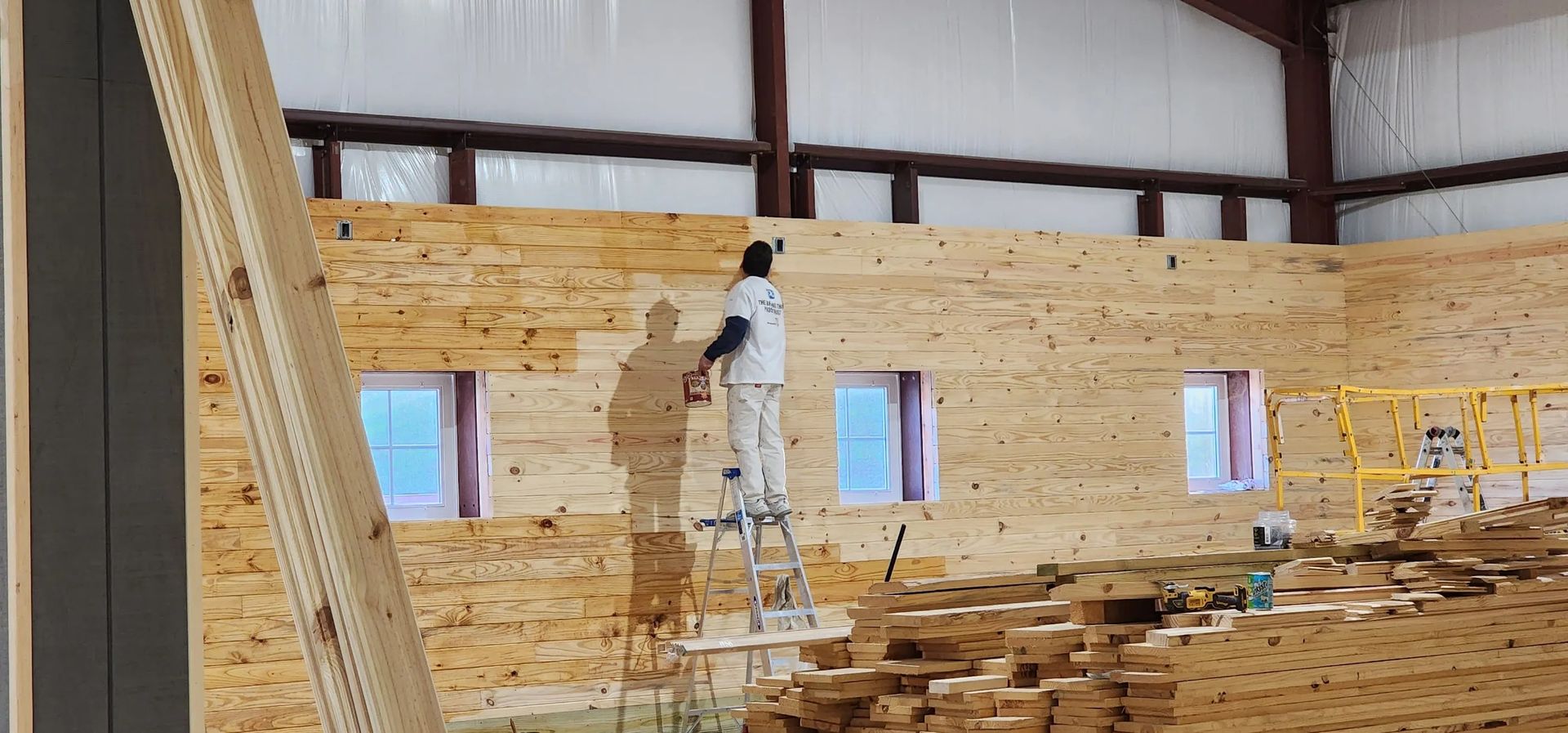 A man is standing on a ladder painting a wooden wall.