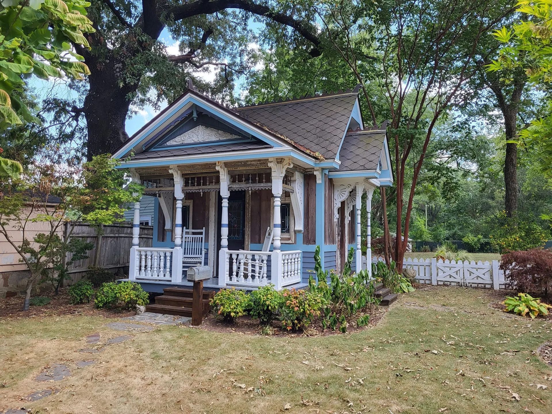A small blue house with a porch in the middle of a yard surrounded by trees.