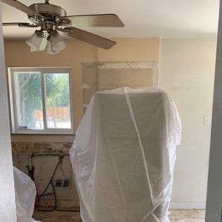 Room with demolition work in progress, kitchen cabinet covered, ceiling fan, window, tan and white walls.