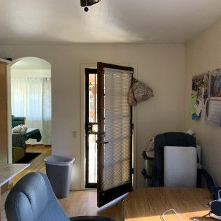 Interior view: Door open to outside, desk, chair, trash can, and archway to another room.