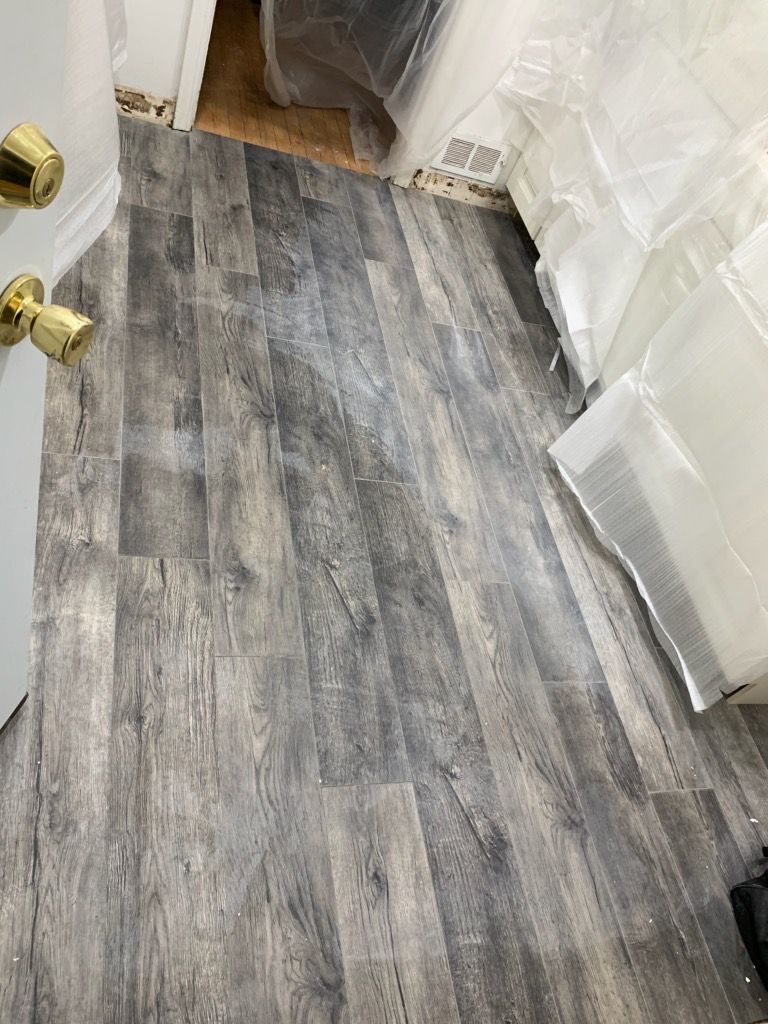 Gray wood-look flooring installed in a room, with a door and stacks of plastic-wrapped items visible.