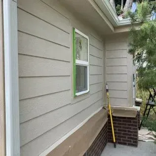 Exterior house painting in progress; tan siding with a white-framed window taped for paint.