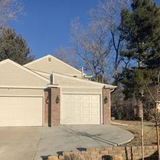 Beige house with two-car garage, brick accents, concrete driveway, and trees under a blue sky.