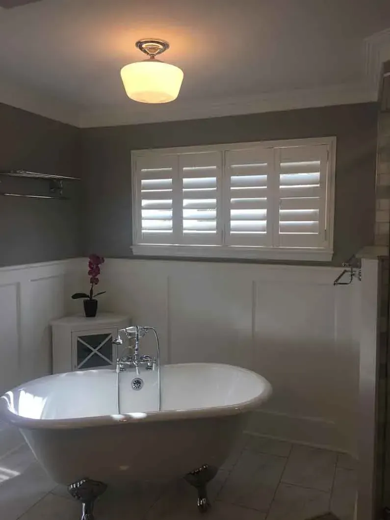 Bathroom with a claw-foot tub, white shutters, and gray walls. A small cabinet and a light fixture are also visible.