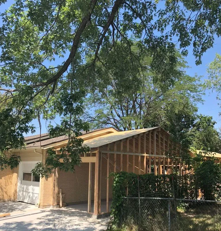 Garage under construction with wooden frame, beige siding, and surrounding trees under a blue sky.