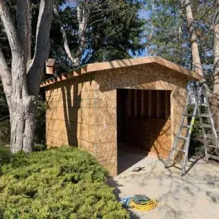 Garage under construction, framed with OSB, next to trees. A ladder is propped nearby.