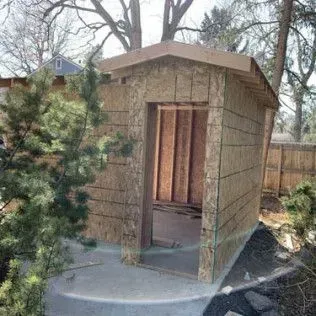 A small, unfinished wooden shed with an open doorway. The shed is outdoors, near bushes and trees.