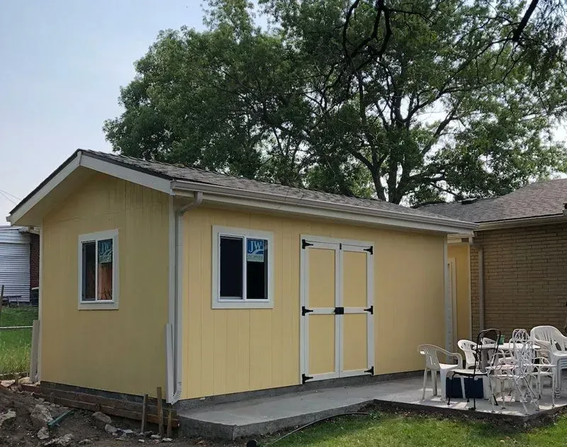 Yellow shed with white trim, windows, and double doors on a concrete patio, with green grass and trees.